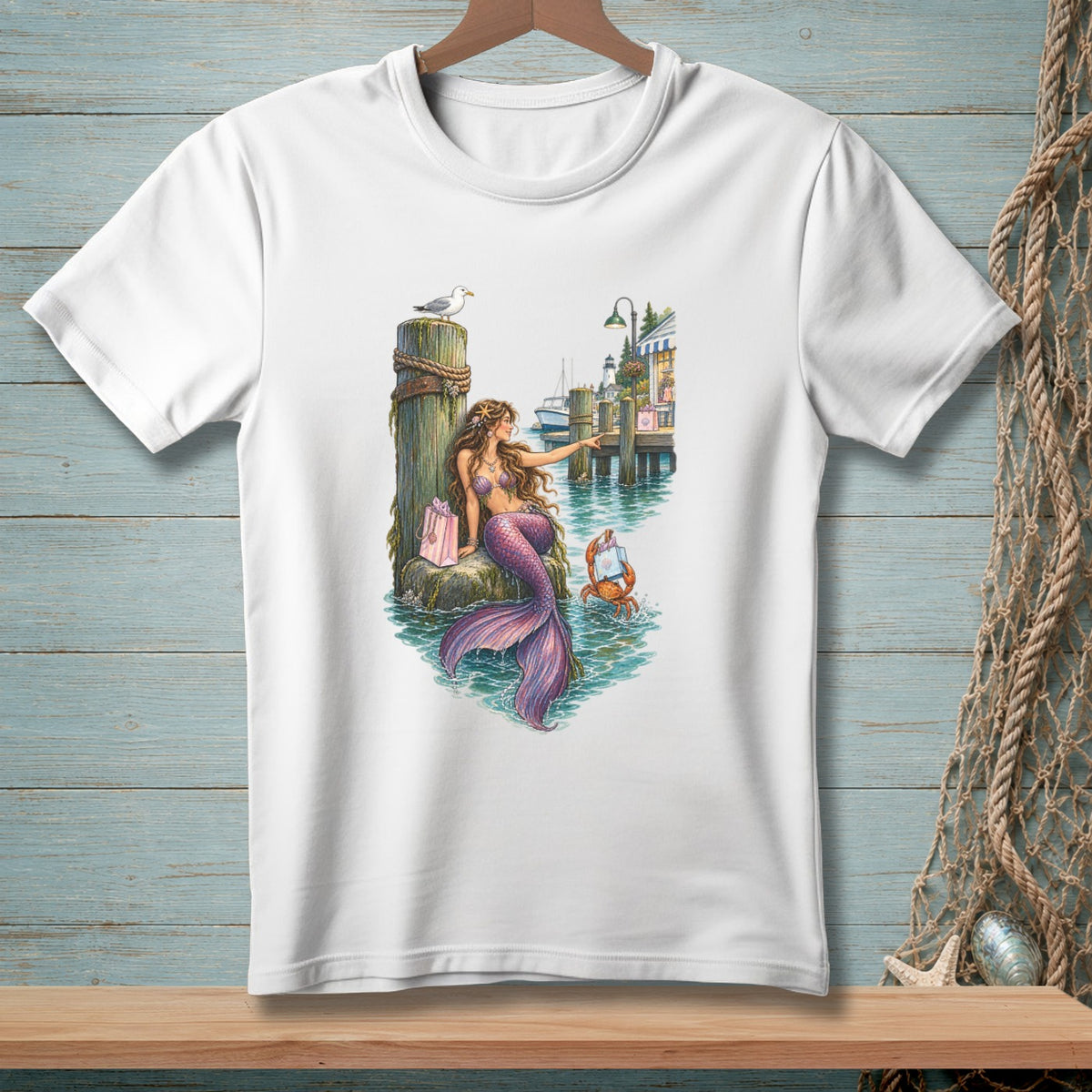 White t-shirt featuring a watercolor illustration of a mermaid by a pier with a crab and shops.