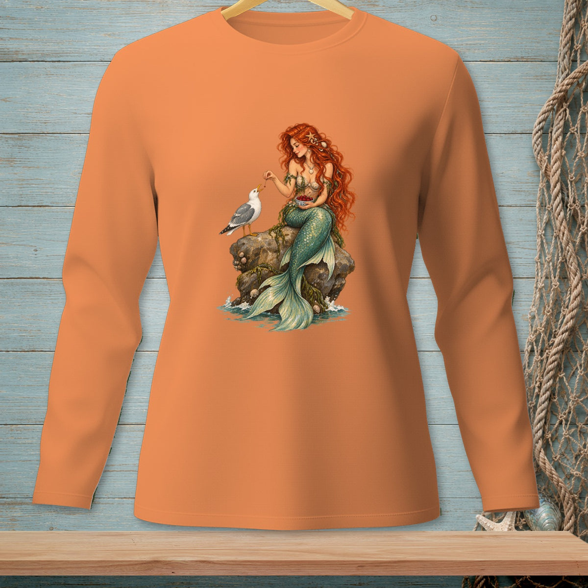 Mermaid with red hair feeding a seagull on a rock, printed on an orange long-sleeve shirt.