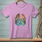 Two mermaids sit on a dock, talking, with the ocean below them.