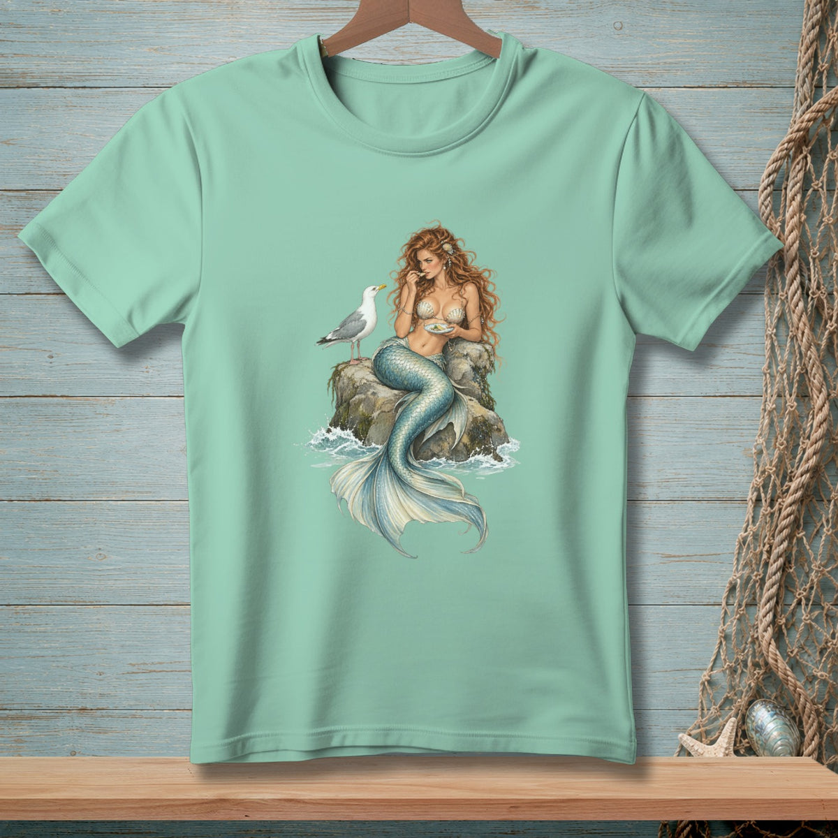 Mermaid with long red hair sits on a rock with a seagull, eating from a plate.