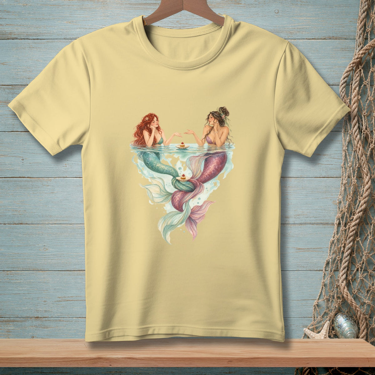 Two mermaids with colorful tails sit at a table in water, serving cake.