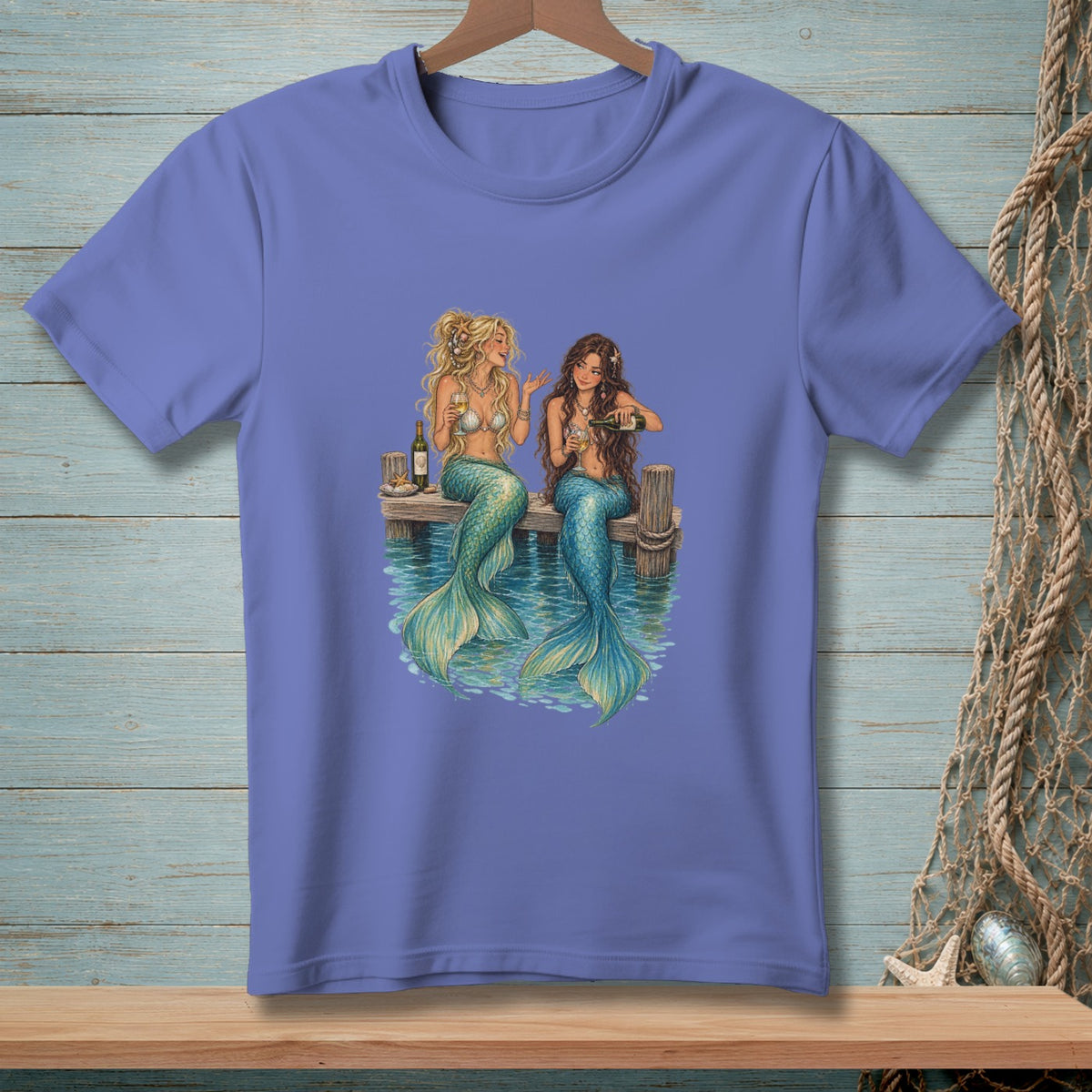 Two mermaids with blue tails sit on a pier, drinking from glasses and a bottle.