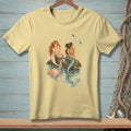 Two mermaids sit on rocks with a seagull flying overhead on a light yellow t-shirt.