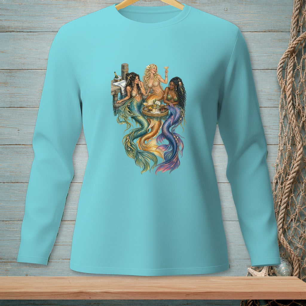 Three mermaids with colorful tails sit at a table on a wooden dock with drinks.