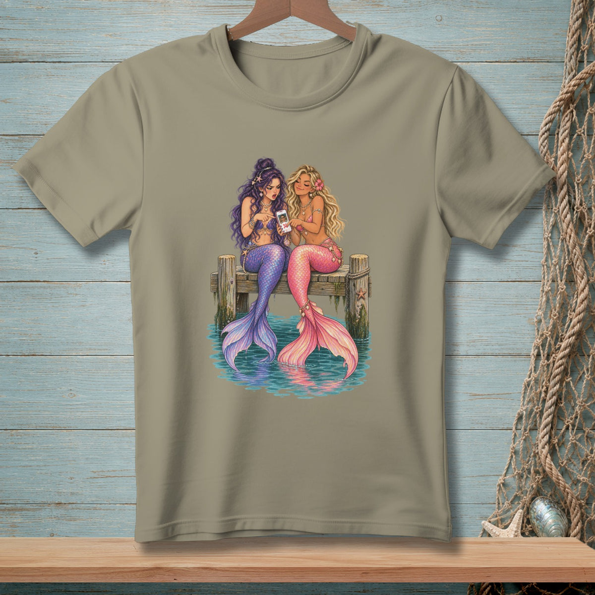 Two mermaids with colorful tails sit on a dock looking at a smartphone.