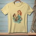 Yellow t-shirt with a graphic of two mermaids by a rock and ocean waves.