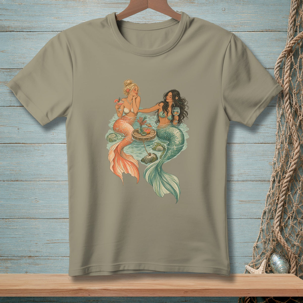 Two mermaids sit in water, one with orange tail, one with teal tail, drinking cocktails.