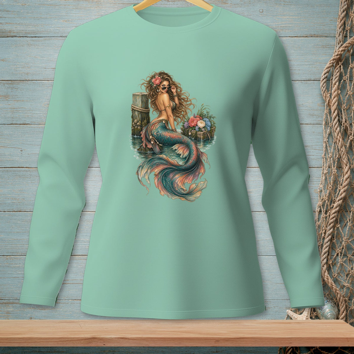 Long sleeve shirt with a mermaid sitting on a pier next to a basket of flowers.