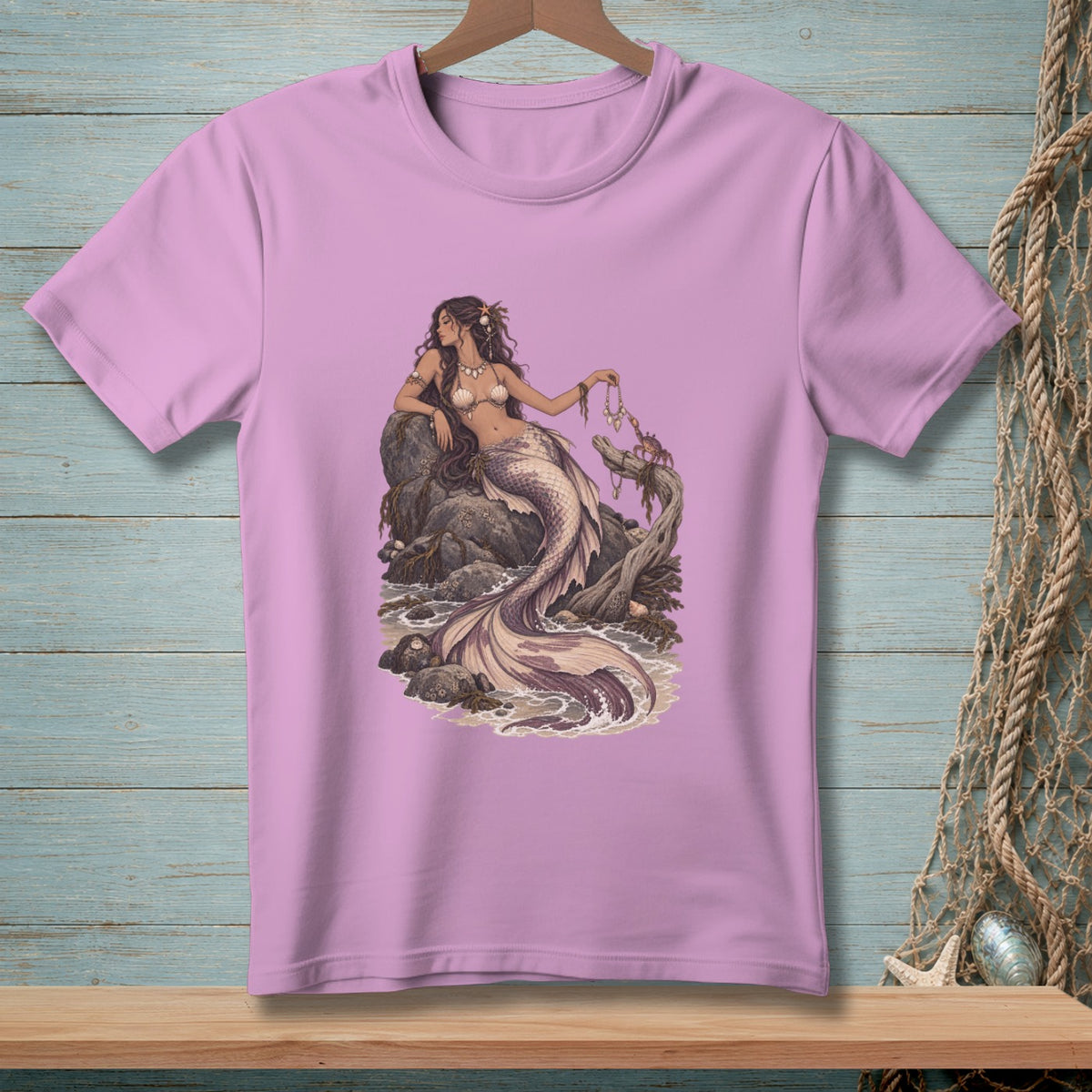 Pink t-shirt with a detailed illustration of a mermaid and a crab on rocks by the sea.