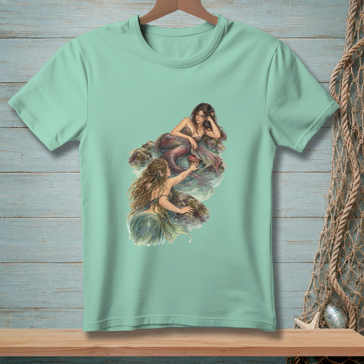 Mint green t-shirt featuring a detailed illustration of two mermaids by the water.