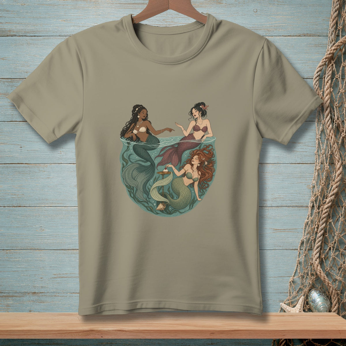 Three mermaids swim together in a circular design on a tan t-shirt.
