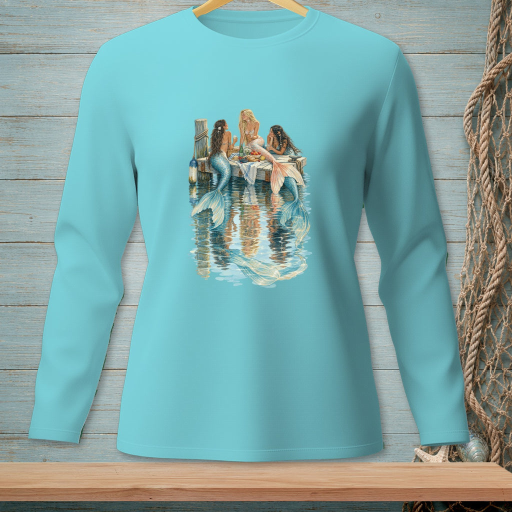 Three mermaids sit on a dock, reflecting in the water.