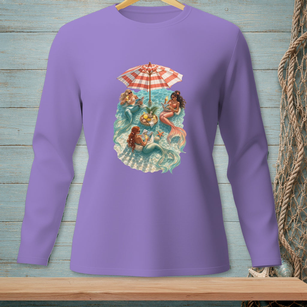 Illustration of mermaids relaxing under an umbrella by the water.