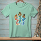 Three mermaids with colorful tails sit around a table, holding drinks.