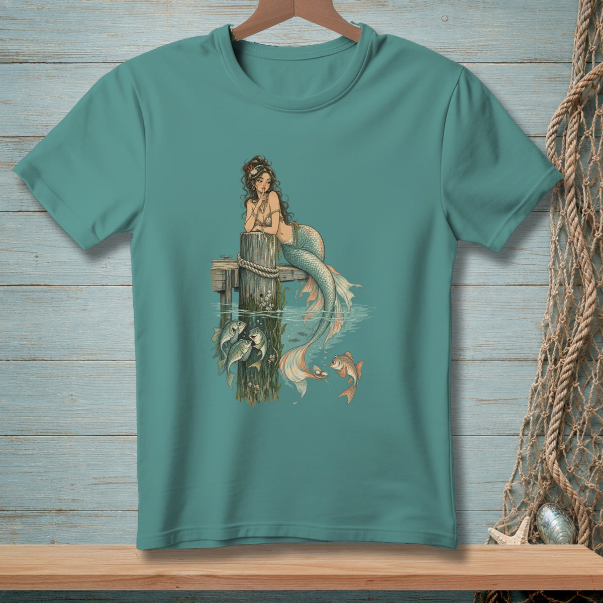 Teal t-shirt with a mermaid sitting on a dock, surrounded by fish.