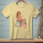 Mermaid with pink tail and crab sits on rock with "Blame the Tail" sign.