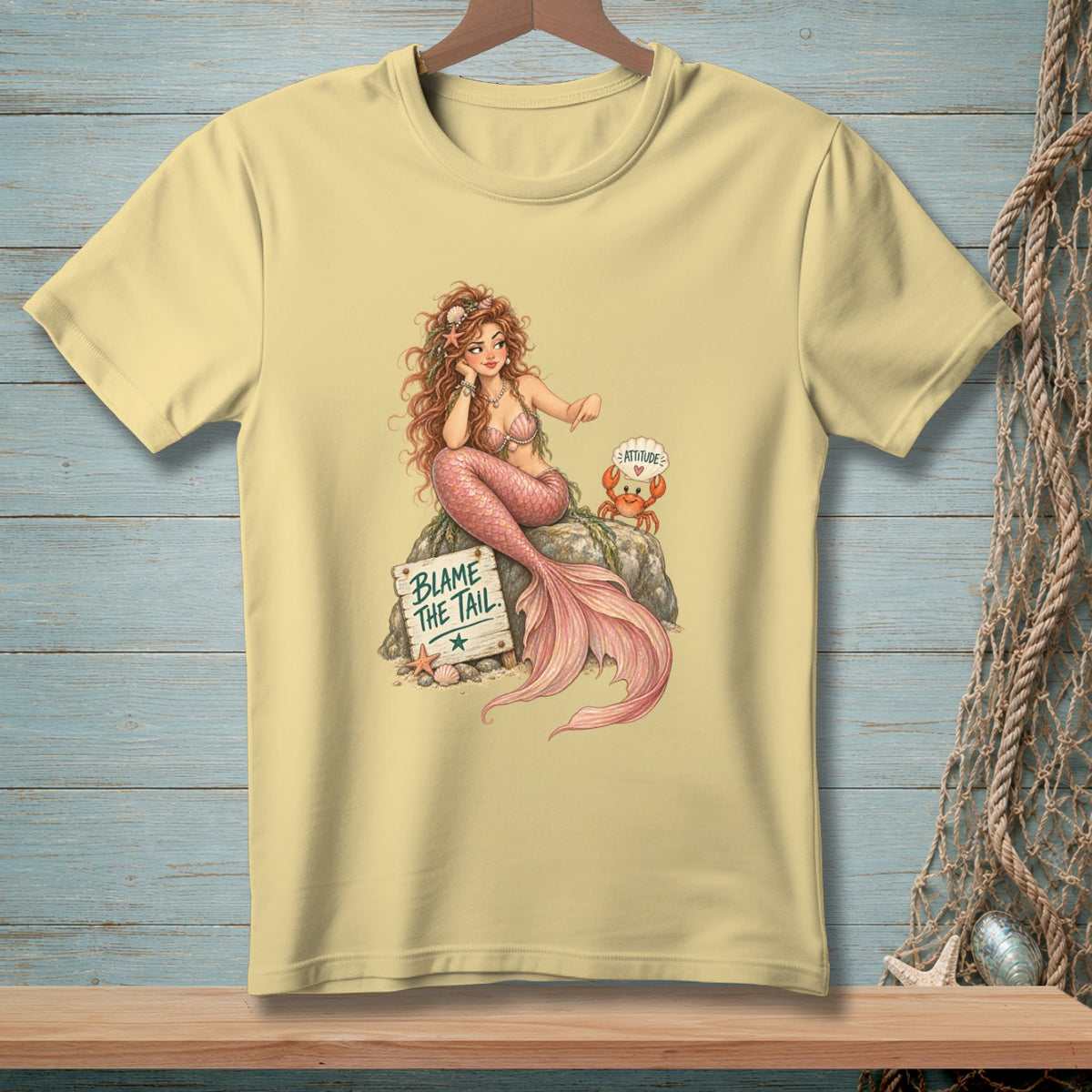 Mermaid with pink tail and crab sits on rock with "Blame the Tail" sign.