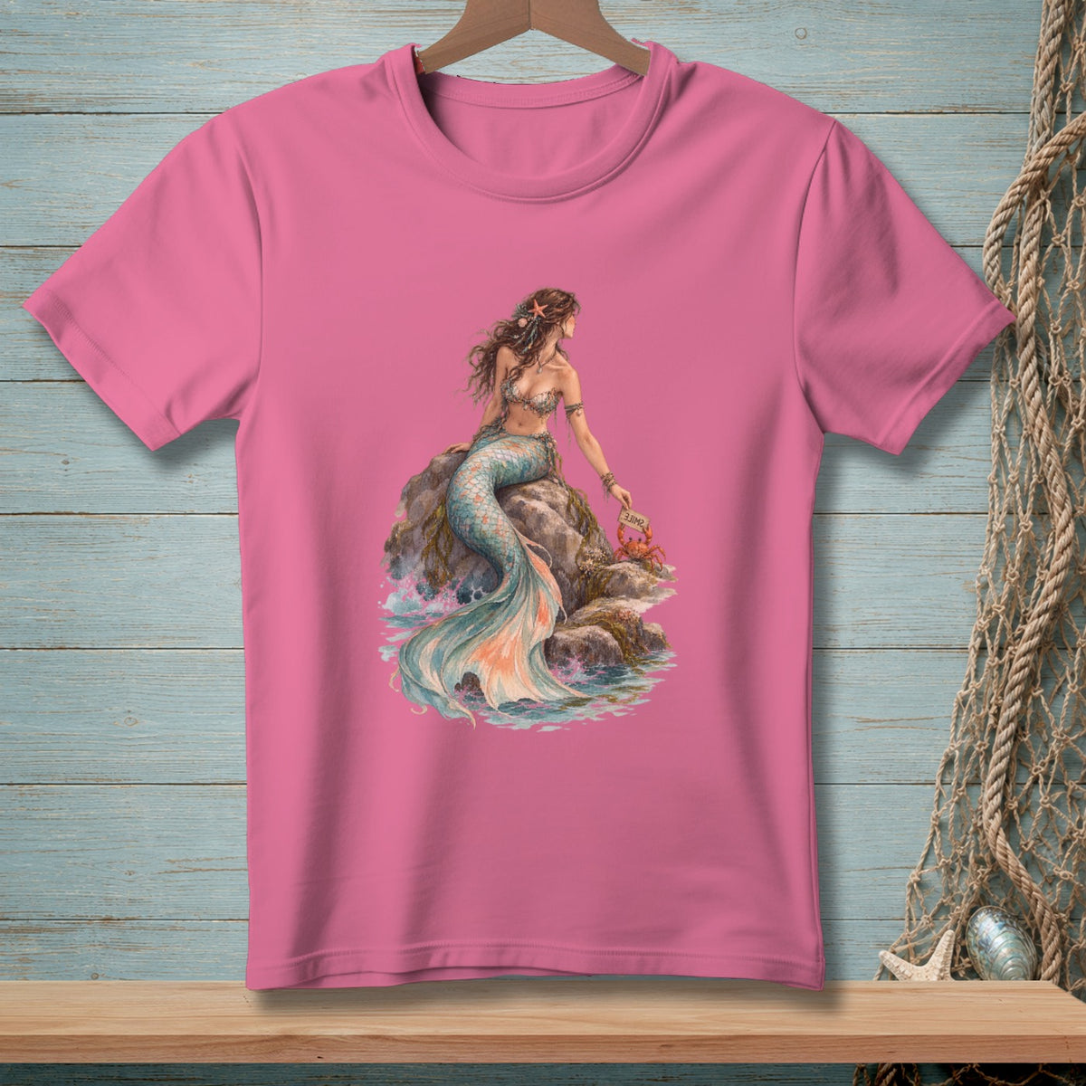 Pink t-shirt with illustration of a mermaid sitting on rocks by the sea with a crab.