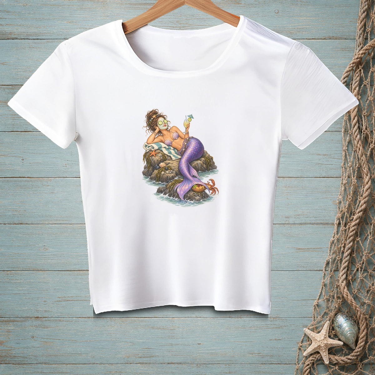 A white t-shirt on a wooden background features a graphic of a mermaid with a face mask relaxing on a rock.