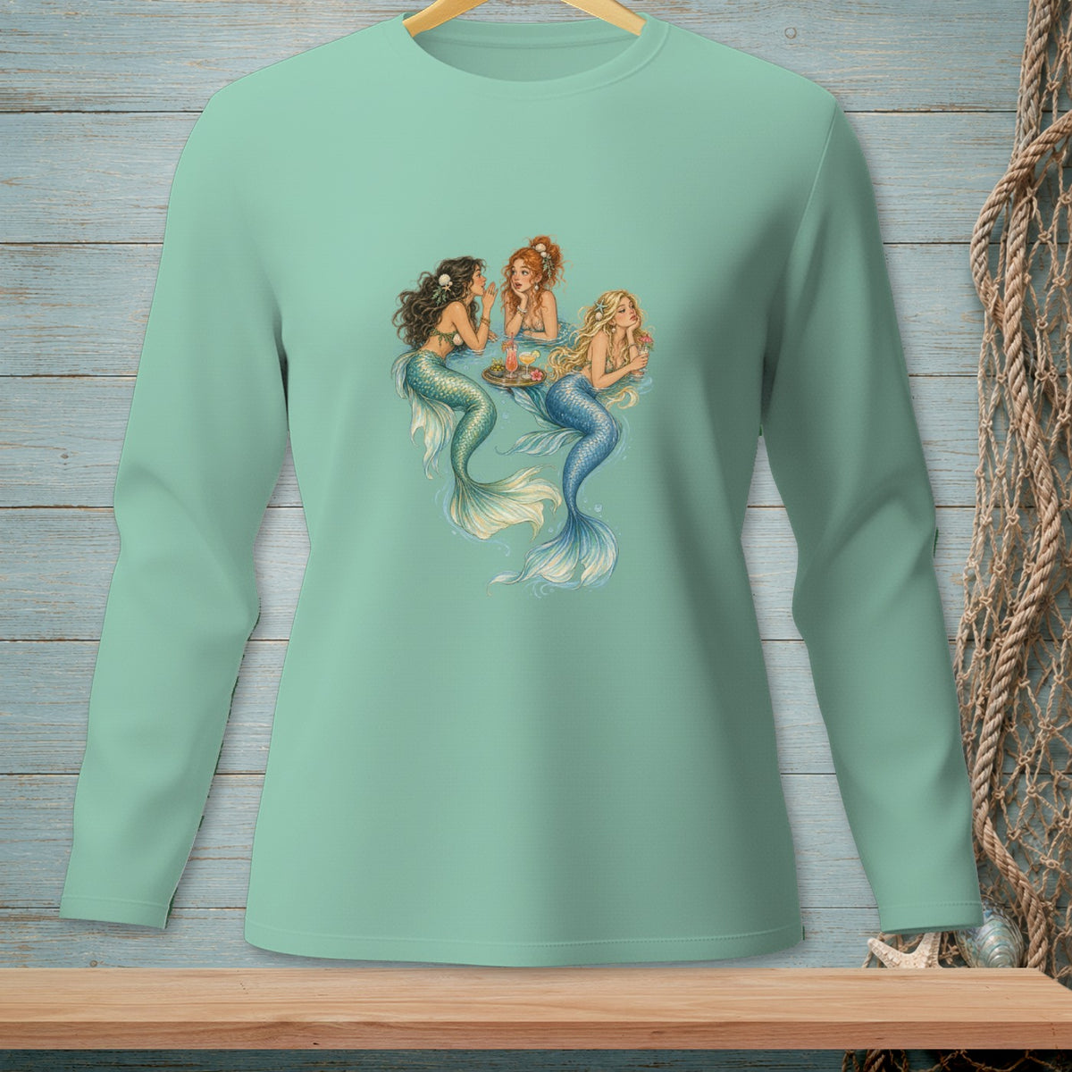 Three mermaids gather around a tray with drinks on a light teal long-sleeve shirt.
