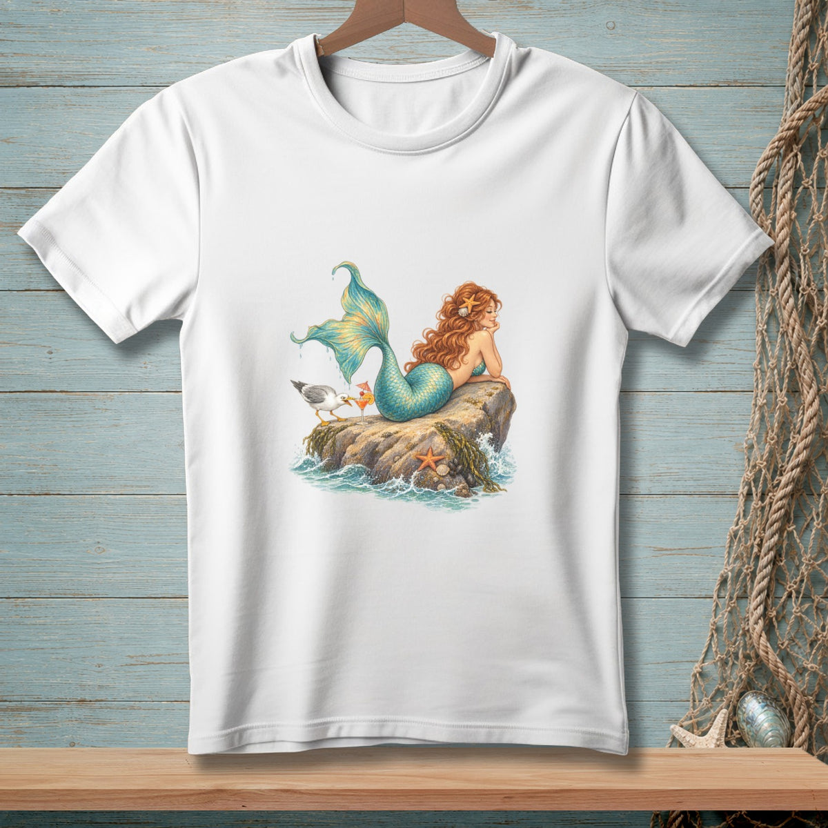 White t-shirt with a watercolor illustration of a mermaid and a seagull on a rock.