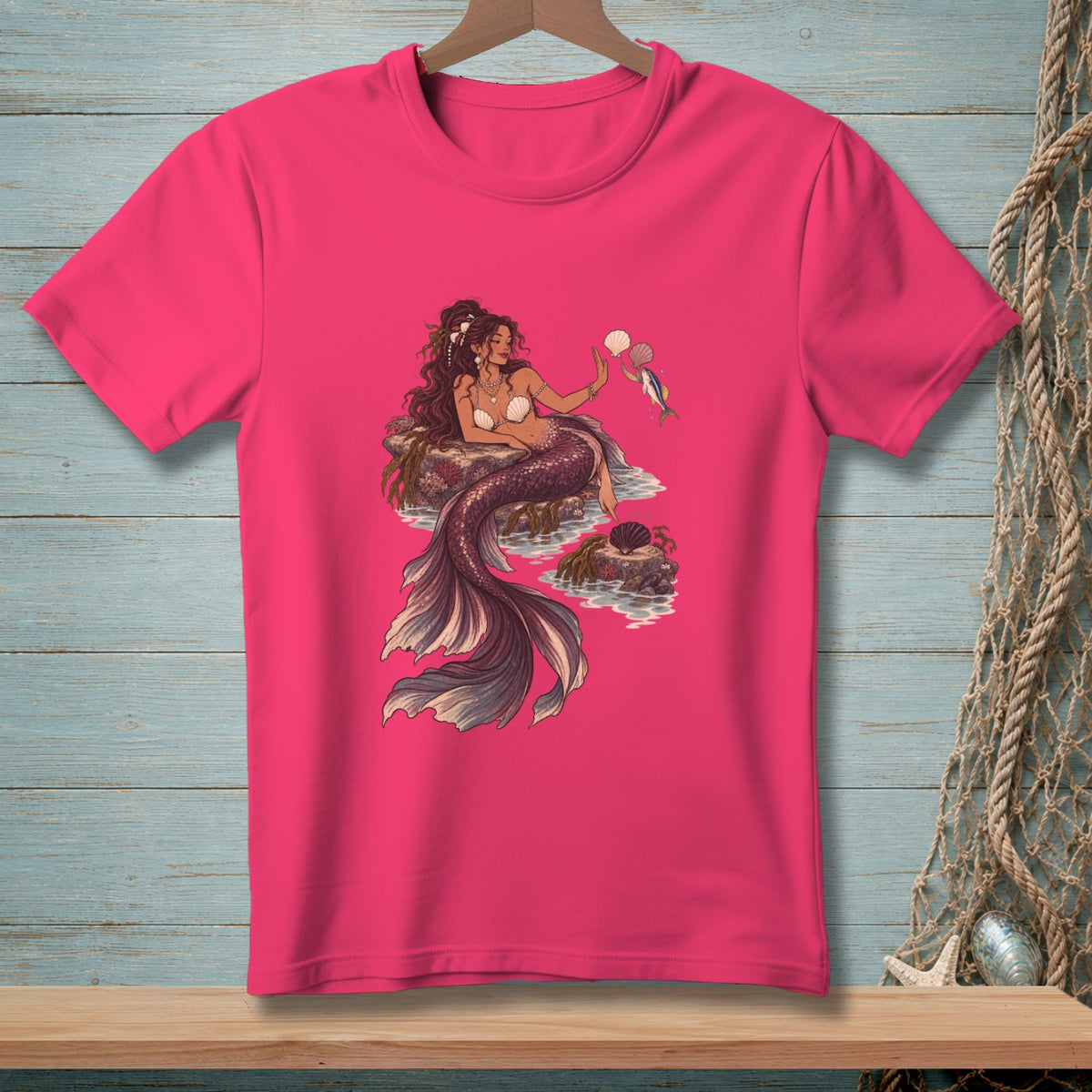 Pink t-shirt with a detailed illustration of a mermaid holding seashells and a fish.