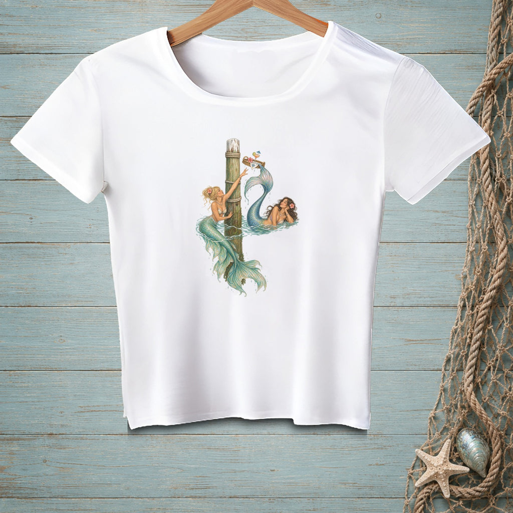 White t-shirt with two mermaids by a wooden post and a small boat.