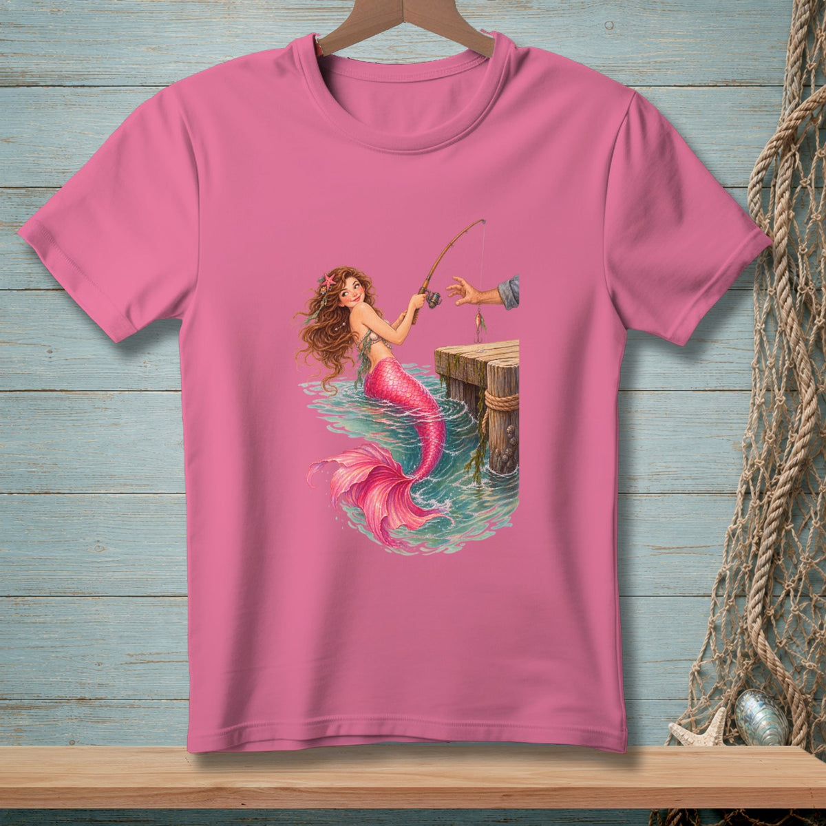 Illustration of a mermaid fishing off a wooden dock on a pink t-shirt.
