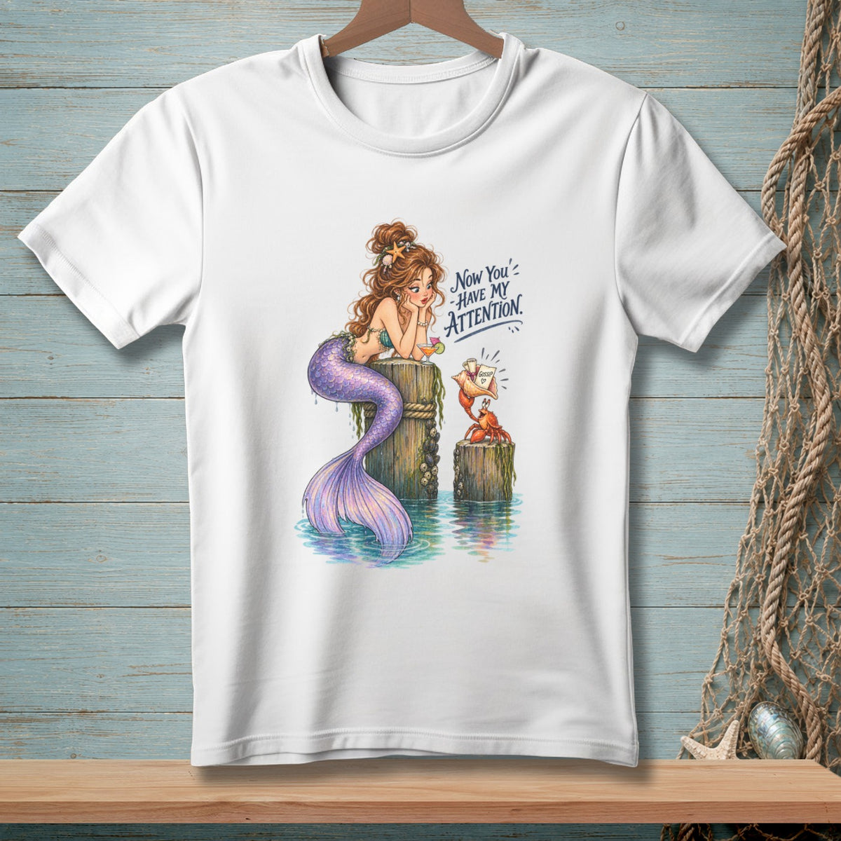 White t-shirt with illustration of a mermaid and crab, text reads "Now You' have my attention."