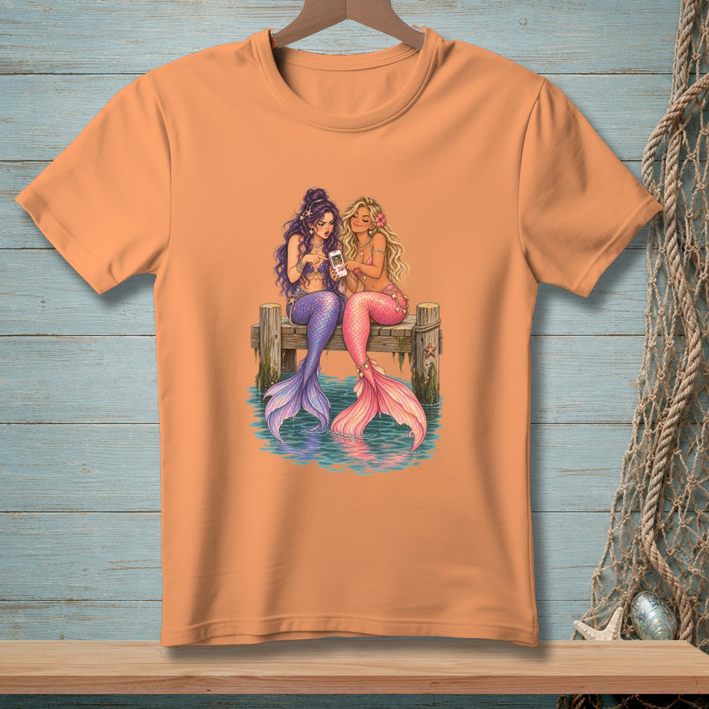 Two mermaids sit on a dock looking at a phone.