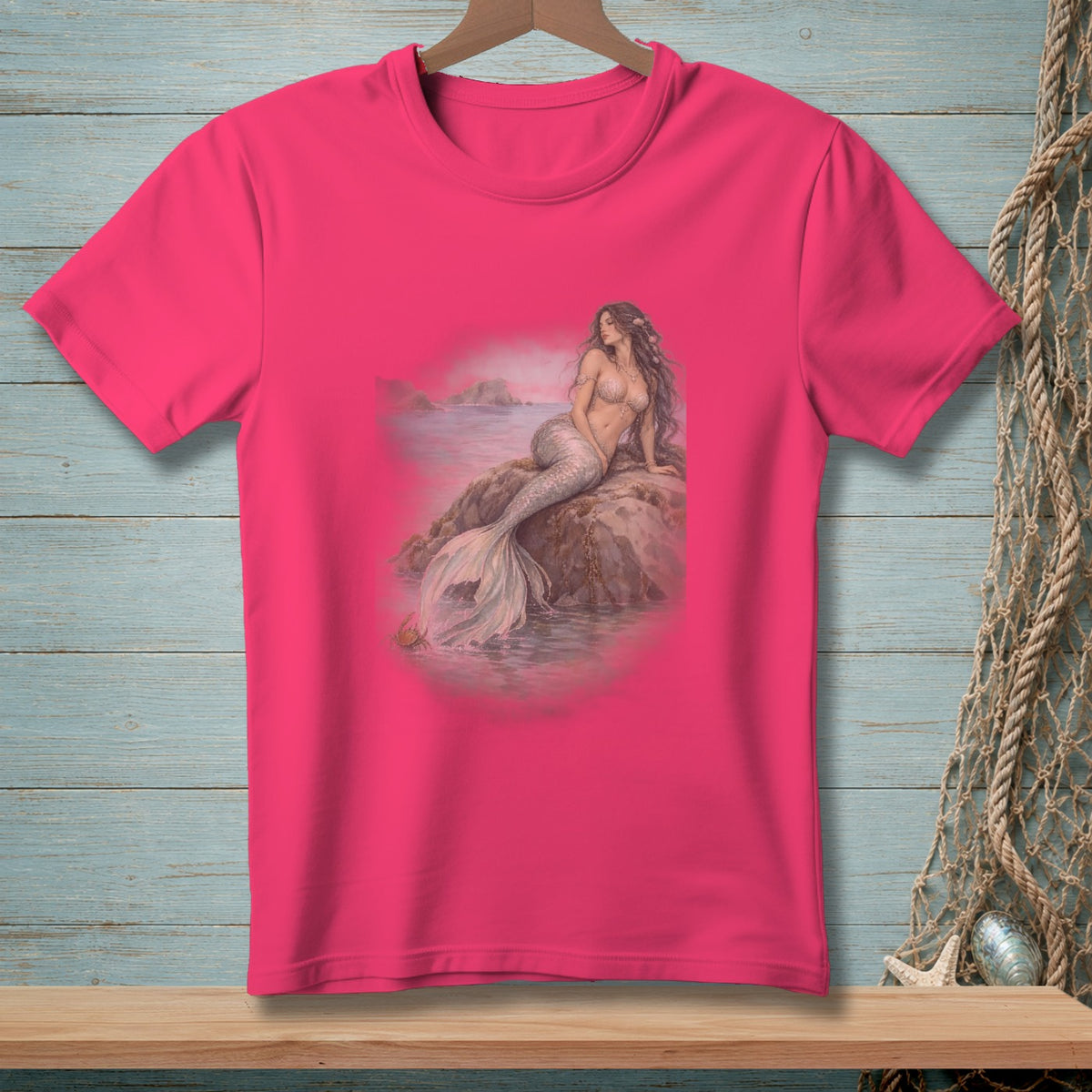 Pink t-shirt with a mermaid sitting on a rock by the water.