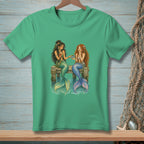 Two mermaids sit on a wooden pier, one offering the other a seashell dish.