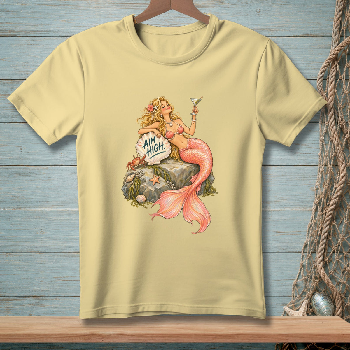 Pale yellow t-shirt with a blonde mermaid holding a cocktail, sitting on a rock with text.