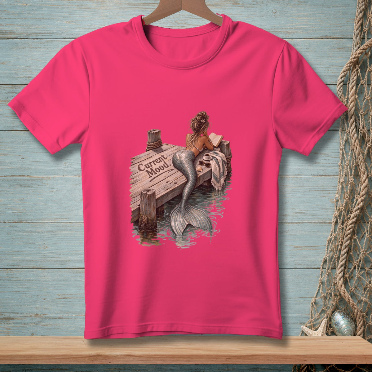 Pink t-shirt with mermaid reading on dock, text "Current Mood."