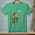 Green t-shirt with a mermaid sitting on a dock, a seagull flies overhead.