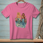 Two mermaids with drinks sit on a dock by the water.