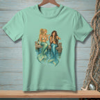Two mermaids with blue and green tails sit on a dock, drinking from wine glasses.