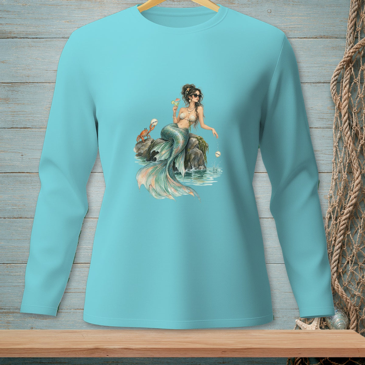 Teal long-sleeve shirt with a mermaid graphic holding a drink on a rock.