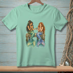 Two mermaids with colorful tails sit on a dock sharing food and drinks.