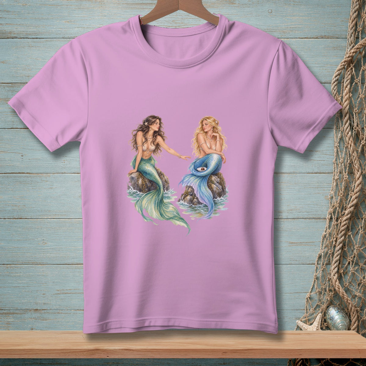 Two mermaids sitting on rocks with waves around them on a pink t-shirt.