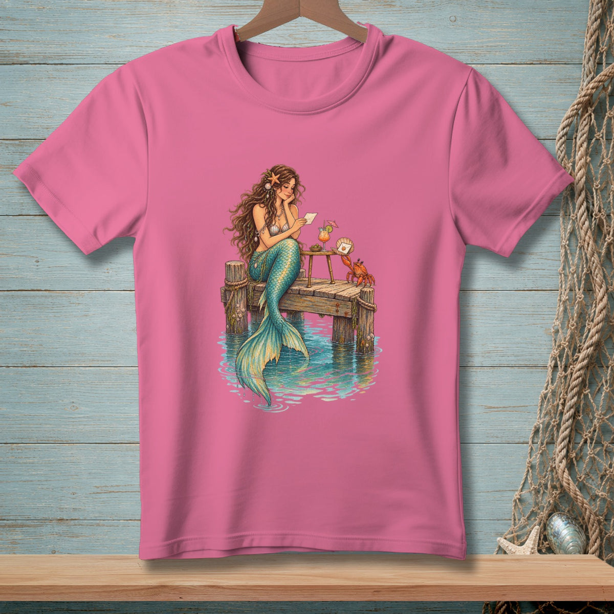 Pink t-shirt with a mermaid sitting on a dock reading a letter with a crab nearby.