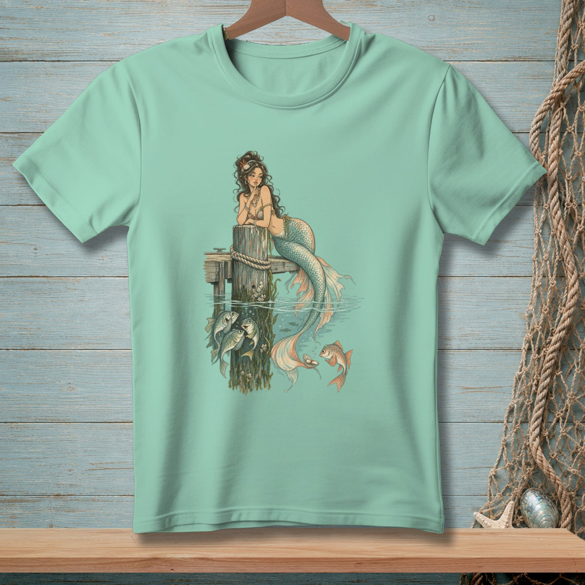 Mint t-shirt with mermaid illustration interacting with fish at a pier.