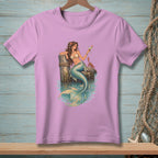 Mermaid with a drink and crab sits on a pier with turquoise water.