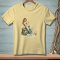 Light yellow t-shirt with a watercolor print of a mermaid holding a drink by the sea.