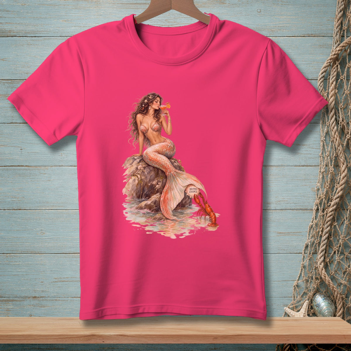 Pink t-shirt with a mermaid sitting on a rock, holding a drink, with a lobster nearby.