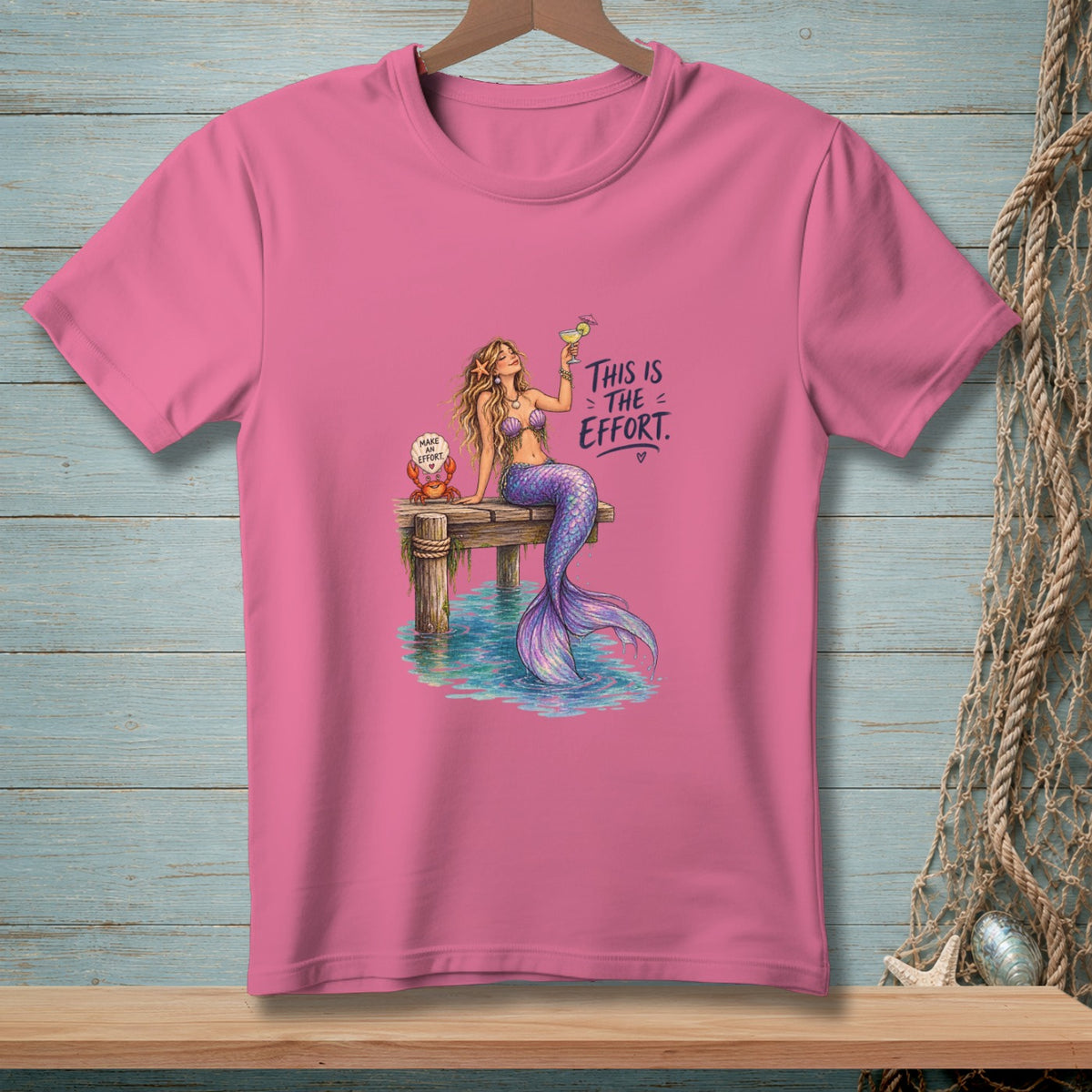Pink t-shirt with mermaid on pier, holding drink, and crab with text.
