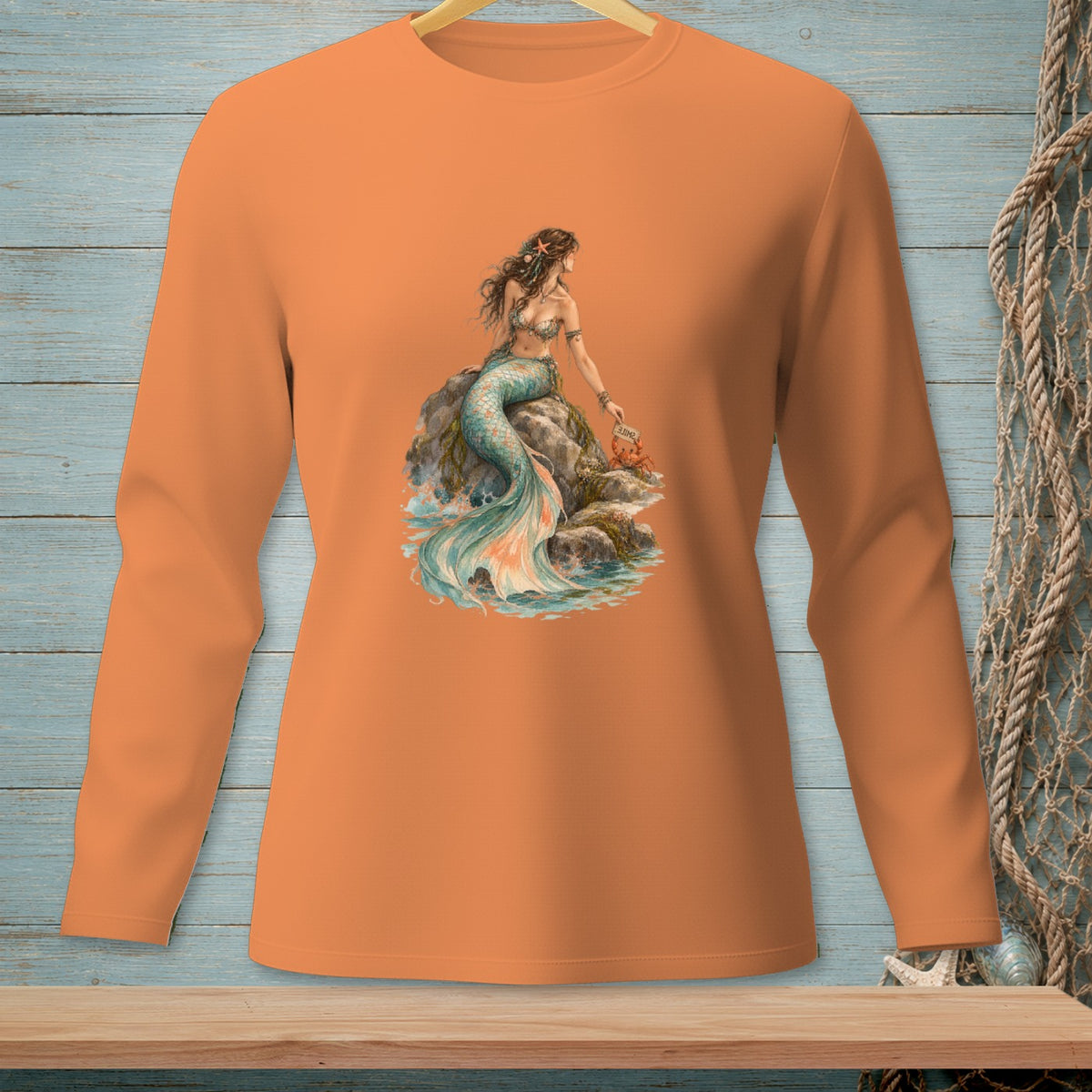 Long-sleeved orange shirt with a watercolor mermaid sitting on rocks by the sea.
