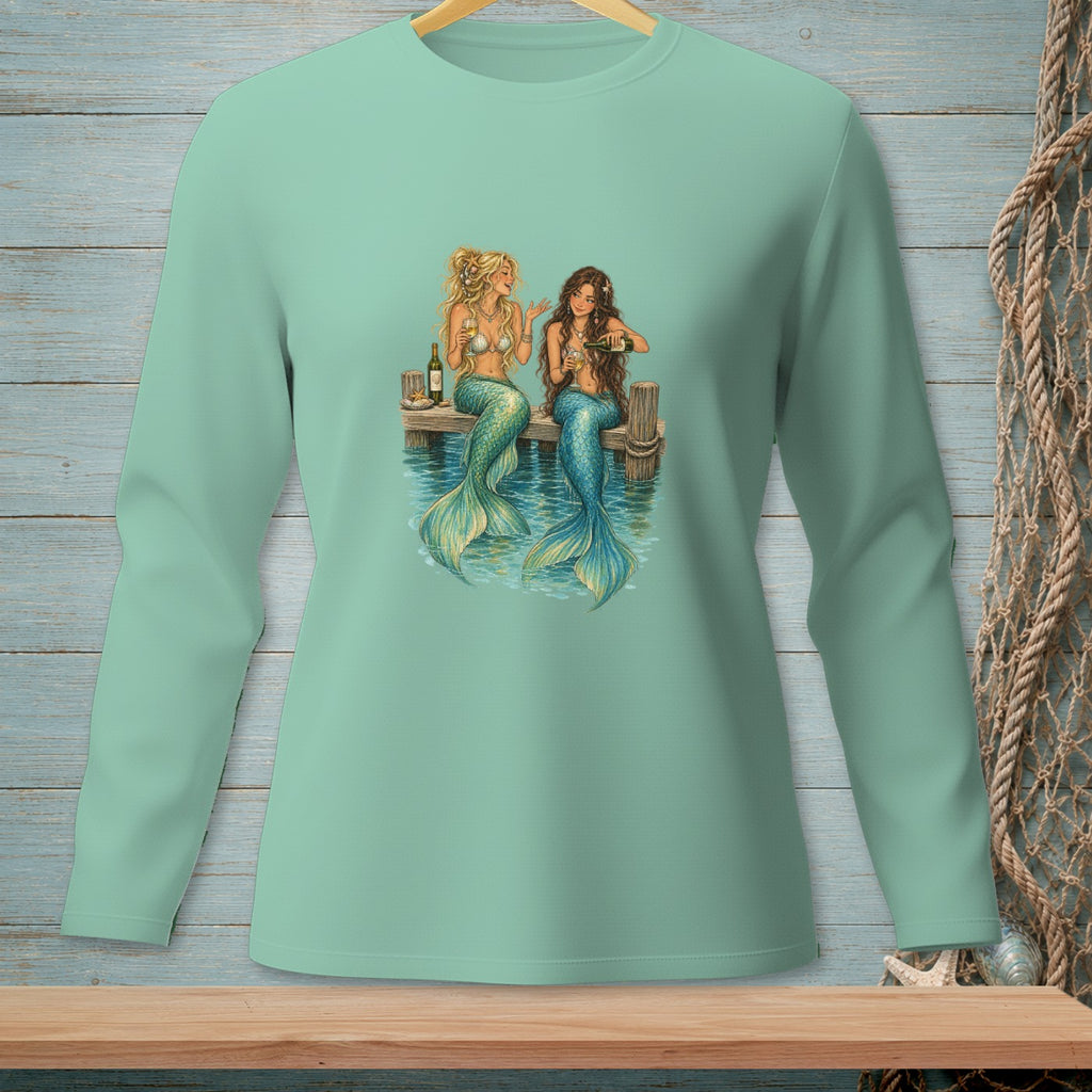Two mermaids with colorful tails sit on a dock, drinking wine.