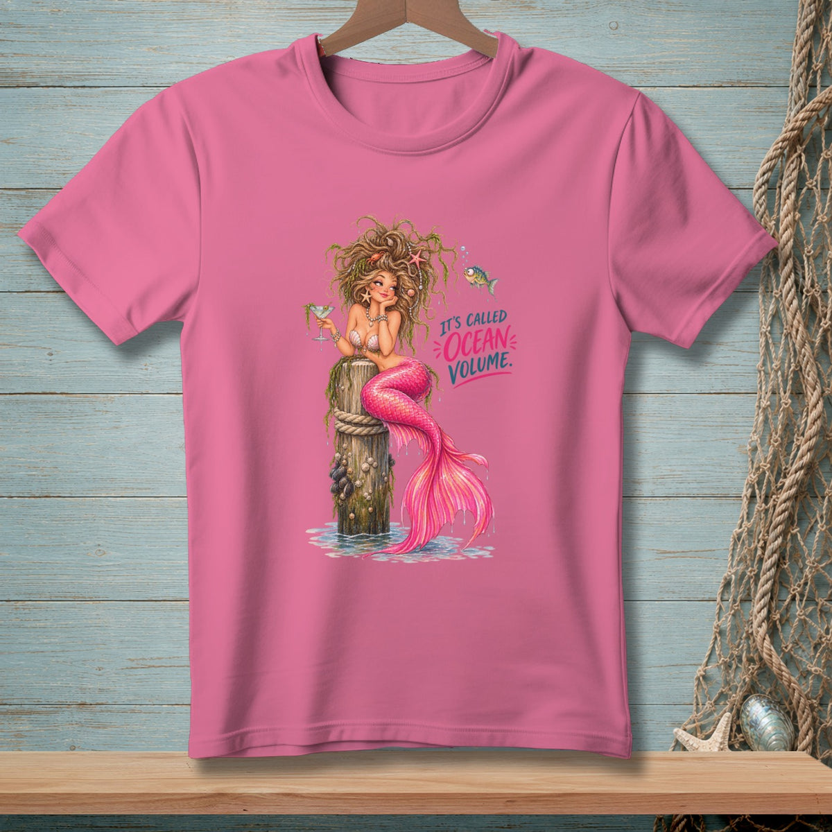 Pink t-shirt with a mermaid sitting on a post, holding a drink. Text says "It's Called Ocean Volume."