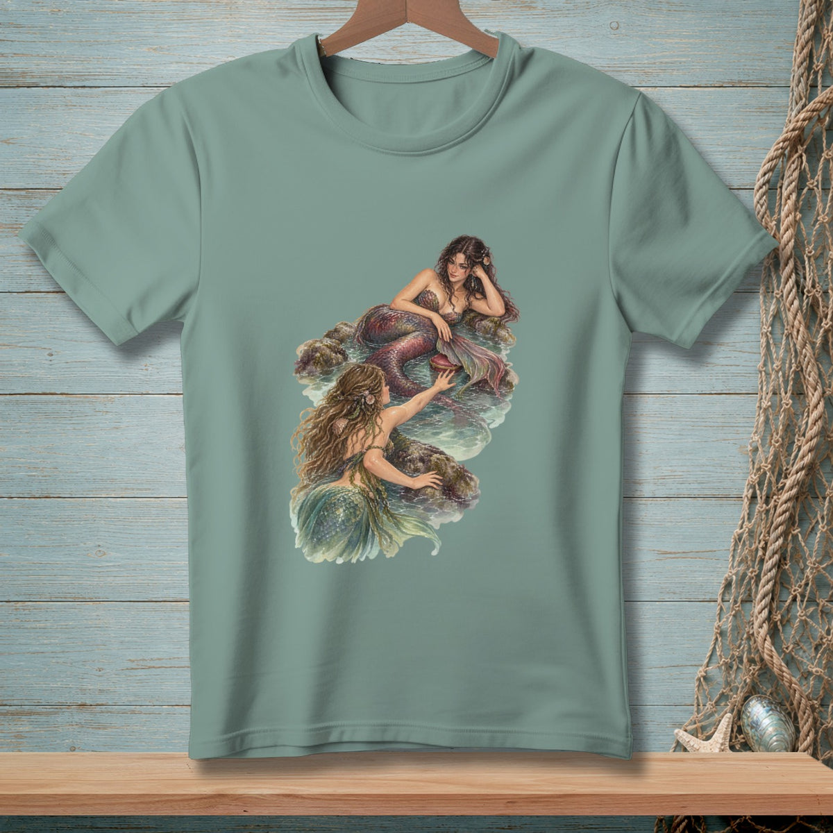 Two mermaids with flowing hair sit on rocks by the water.